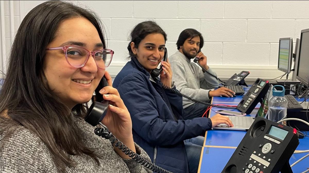 Three students (two female & one male) holding land phones to thier ears with computers in front of them