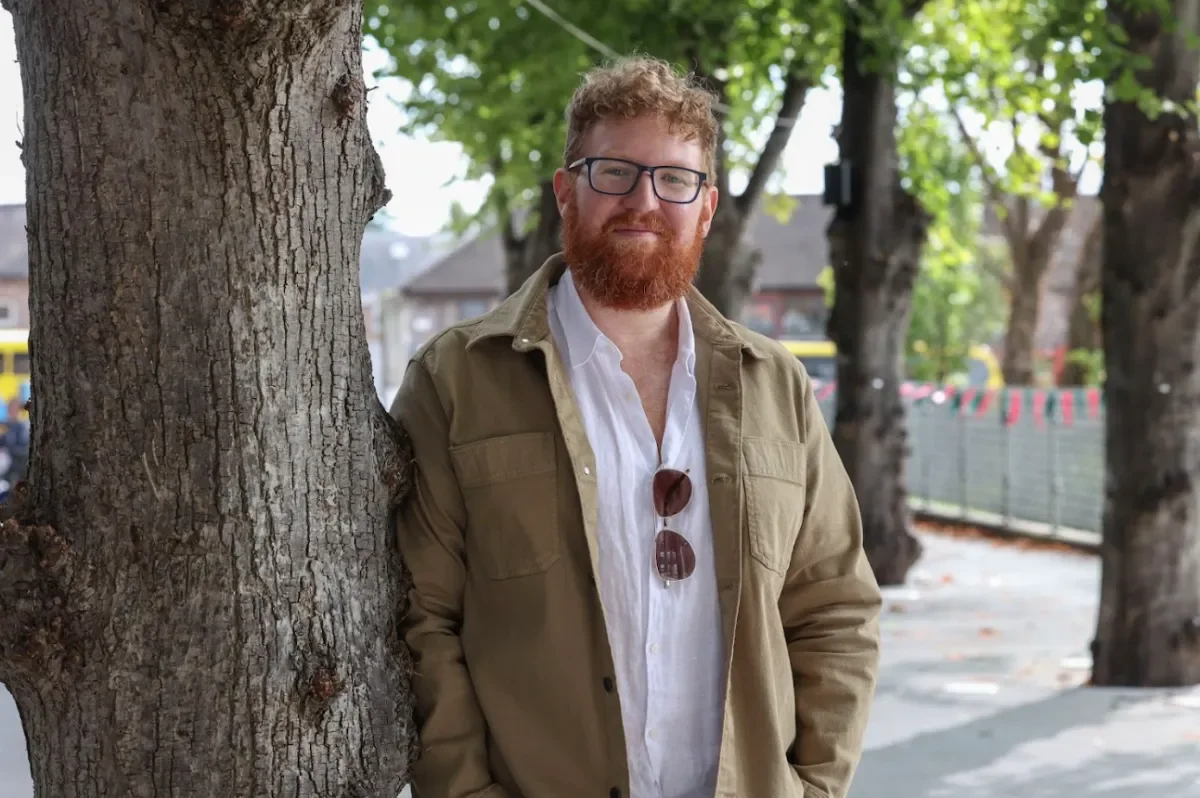Author Dave Rudden who is smiling wears glasses, white shirt and green jumper leans against a tree