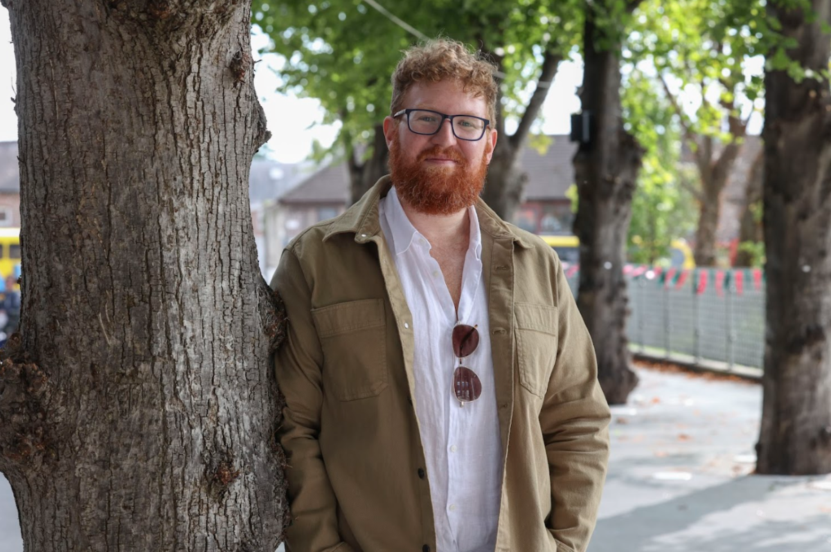 Author Dave Rudden who is smiling wears glasses, white shirt and green jumper leans against a tree 