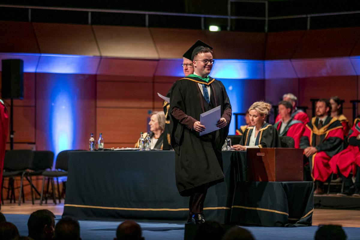 Fergal McGuirk, wearing a graduation cap and gown, walks smiling off the stage after receiving his degree certificate.
