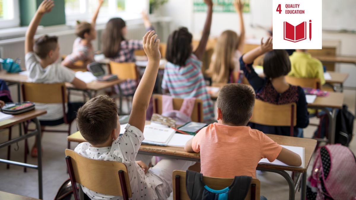 Shows children in class raising their hands to answer a question, in the background the teacher is visible in soft focus