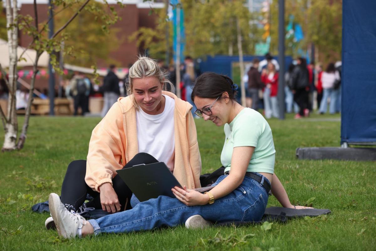 A photo of two young students sitting on some grass, reading from a laptop.