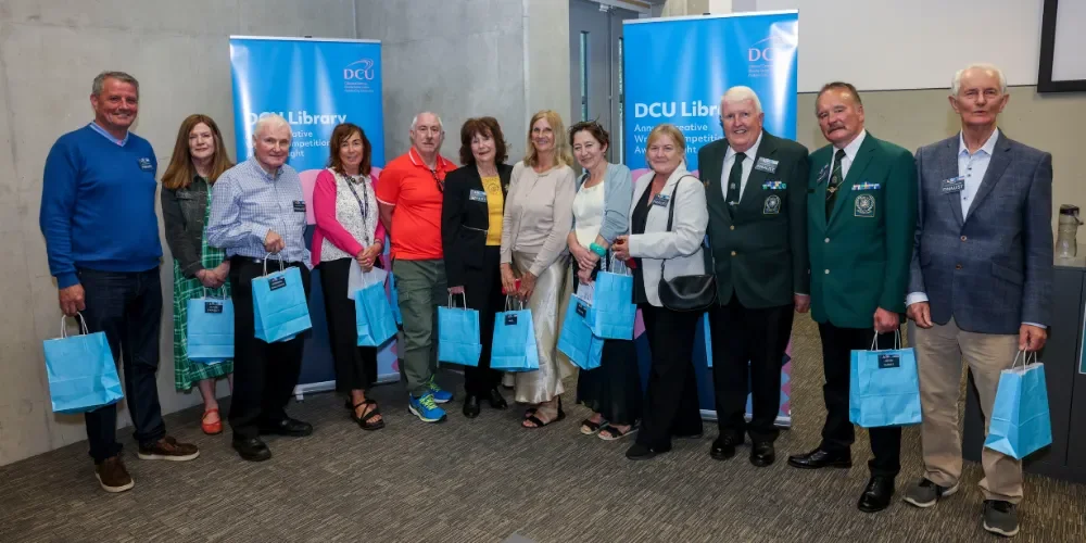 Twelve people holding blue bags facing the camera. Two banners behind the group have DCU Library printed on them.