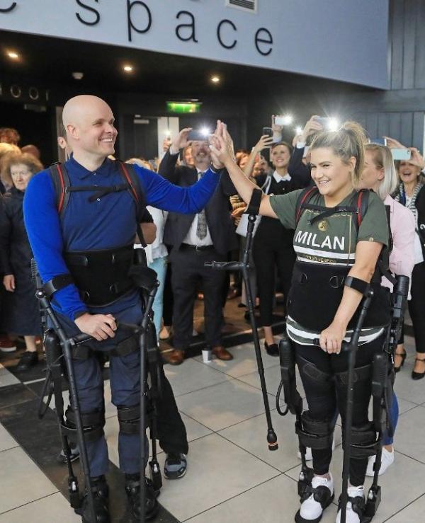 Man and woman in robotic exoskeletons stand in room of people