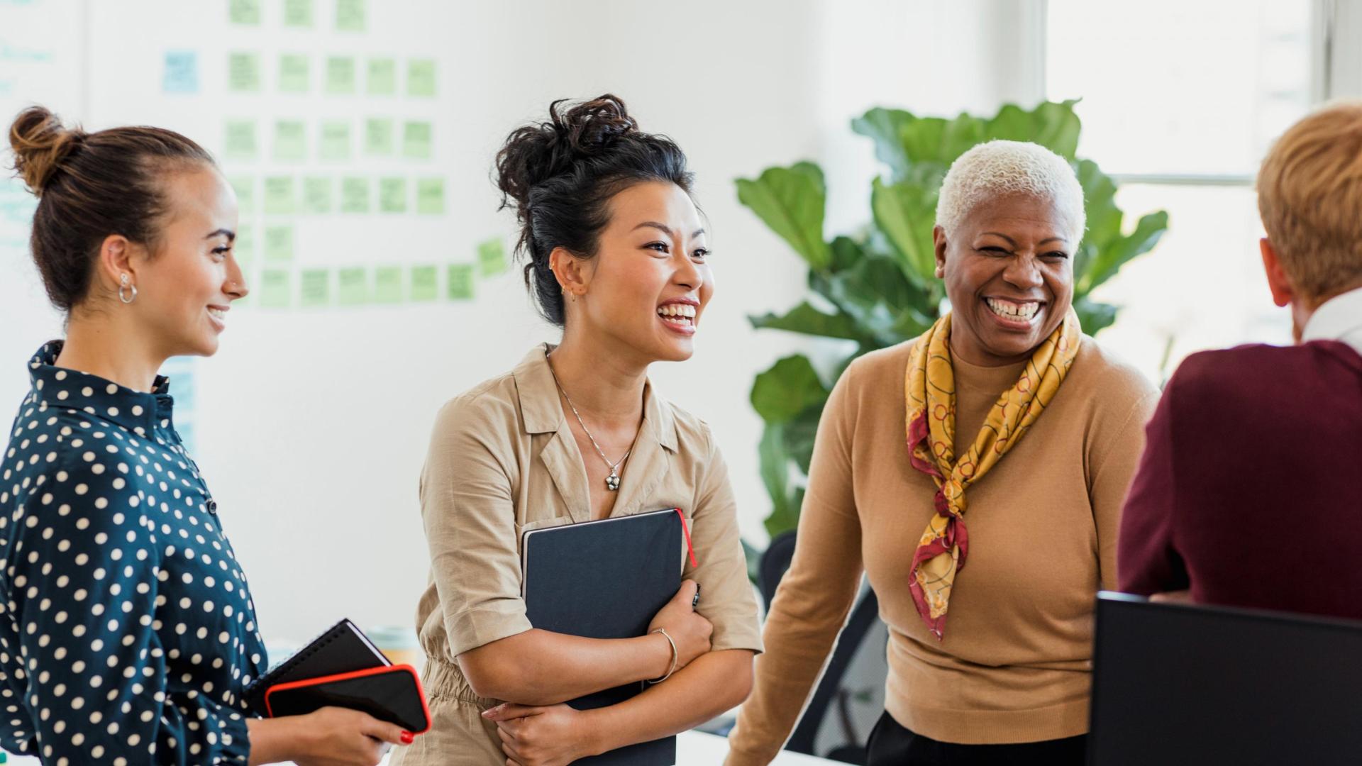 Three women in an office smiling and talking to a male colleague who is facing away from the camera.. 