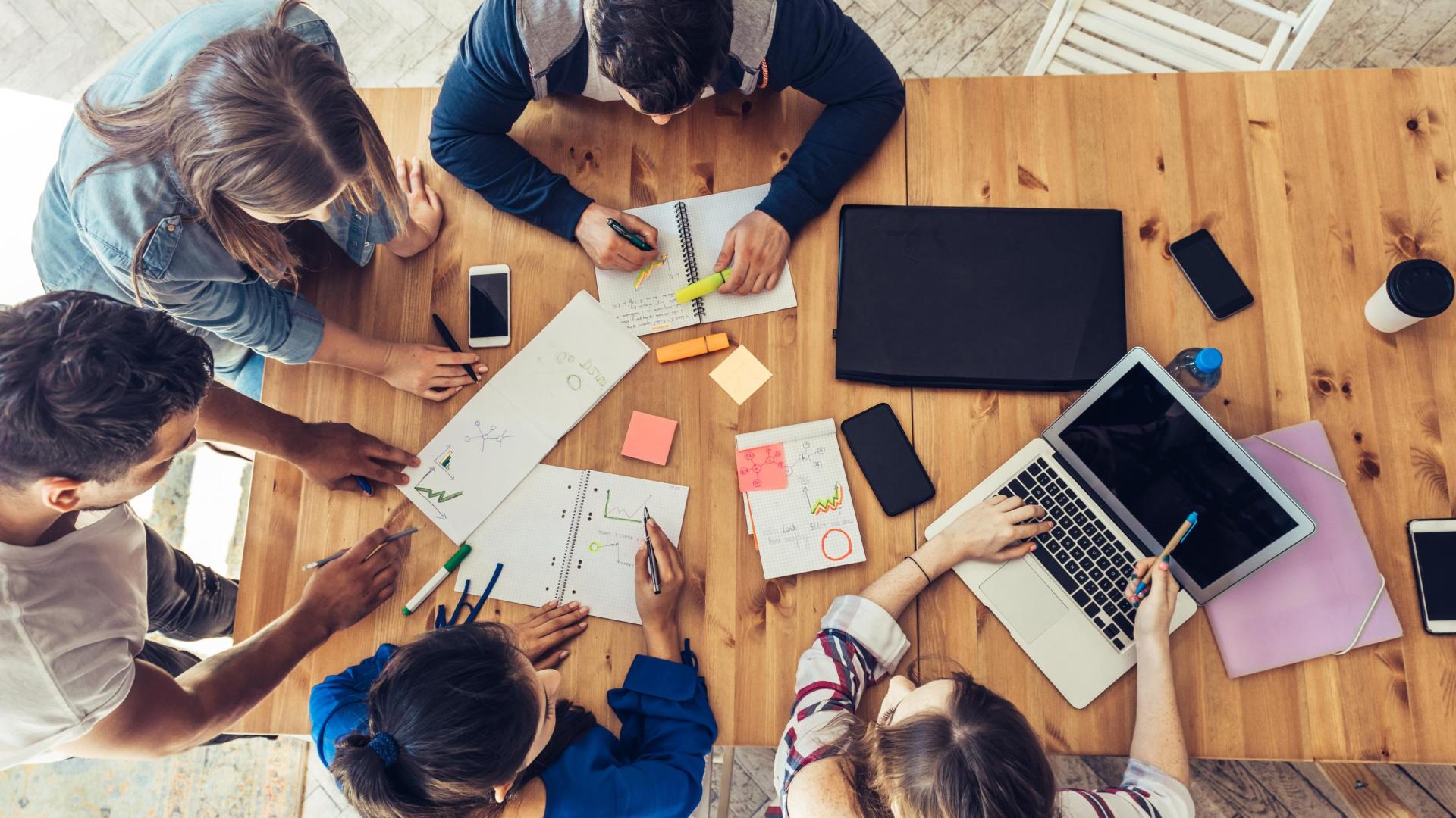 Three females and two males working together with notebooks, markers and a laptop, around a wooden table viewed from above. 