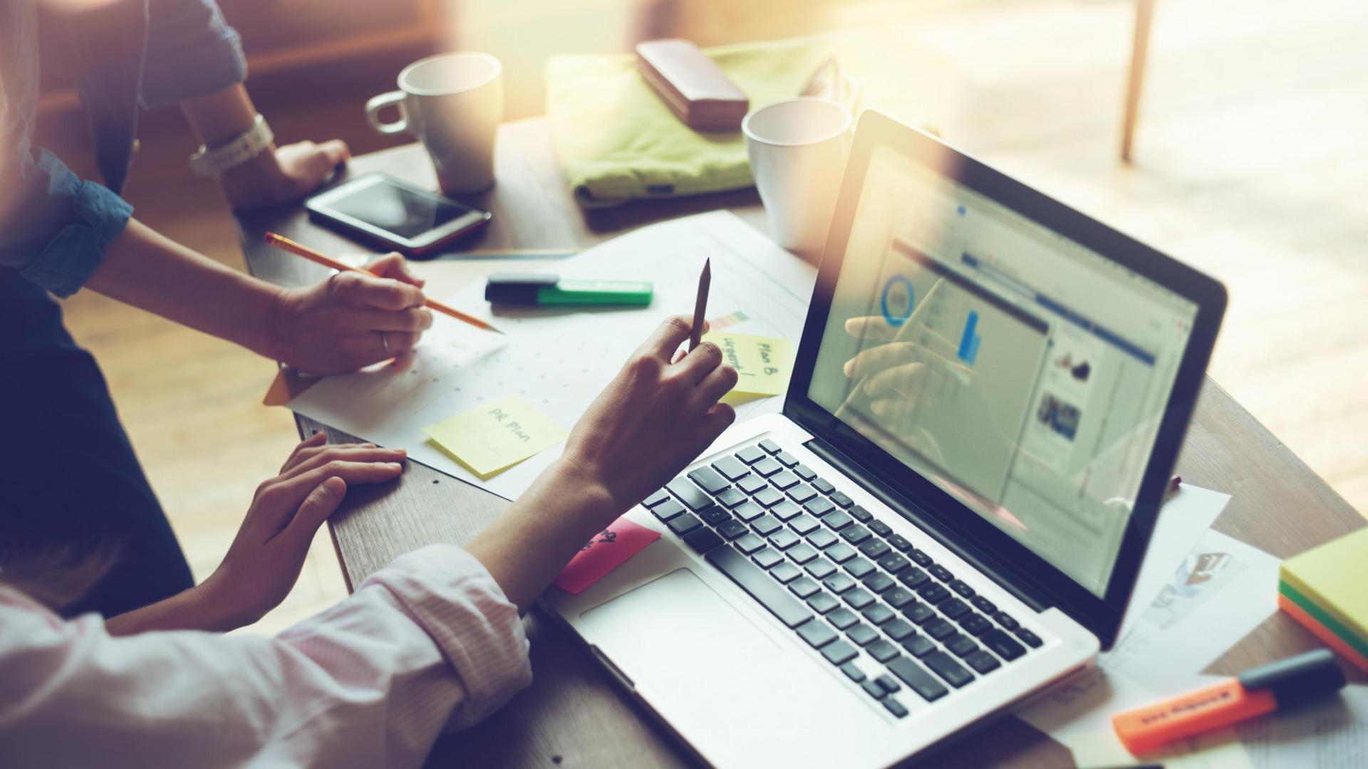 Two people holding pencils working over sheets and an open laptop