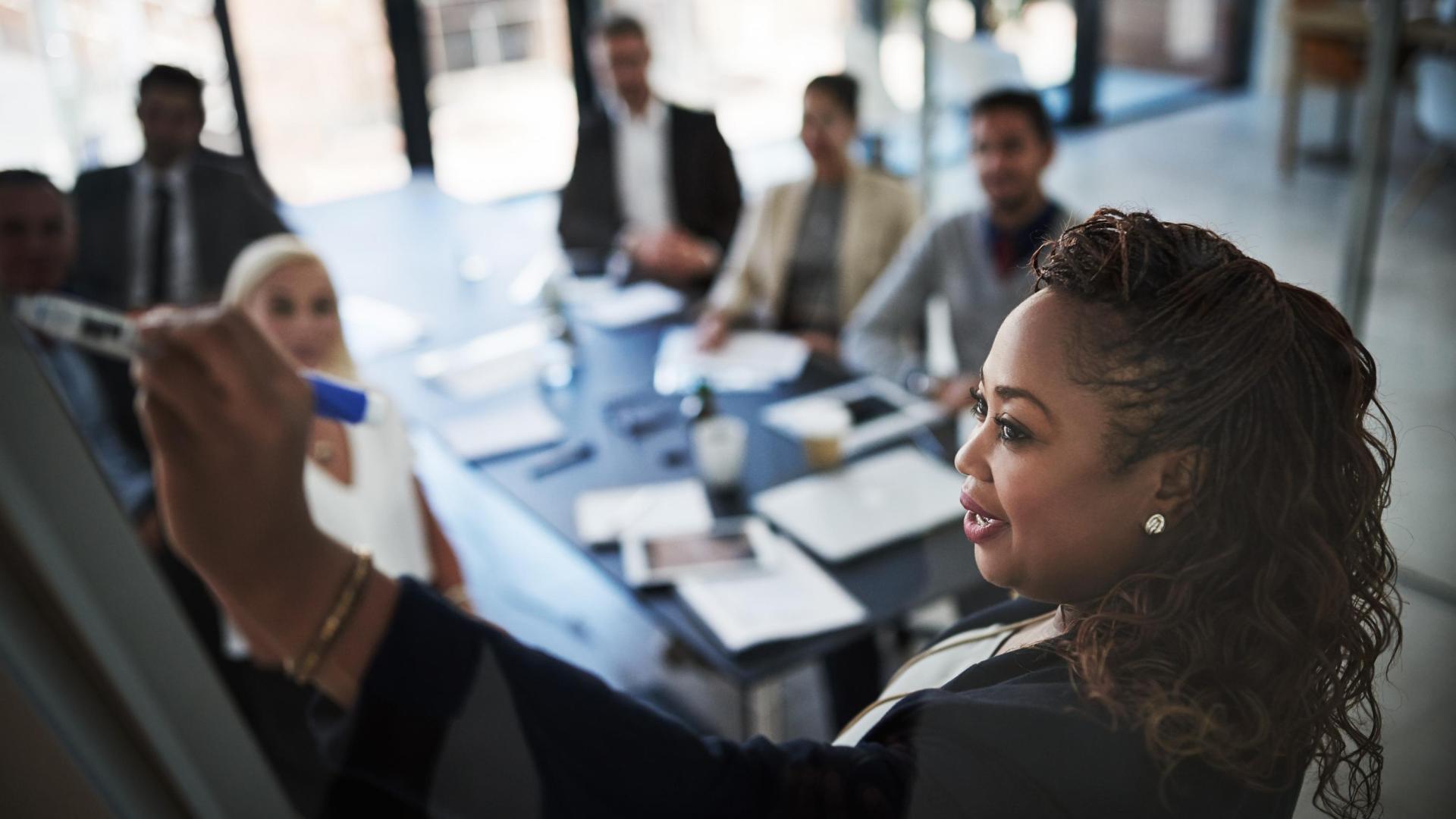 A black woman writing on flipchart paper with a group of colleagues sitting behind her at a table. 