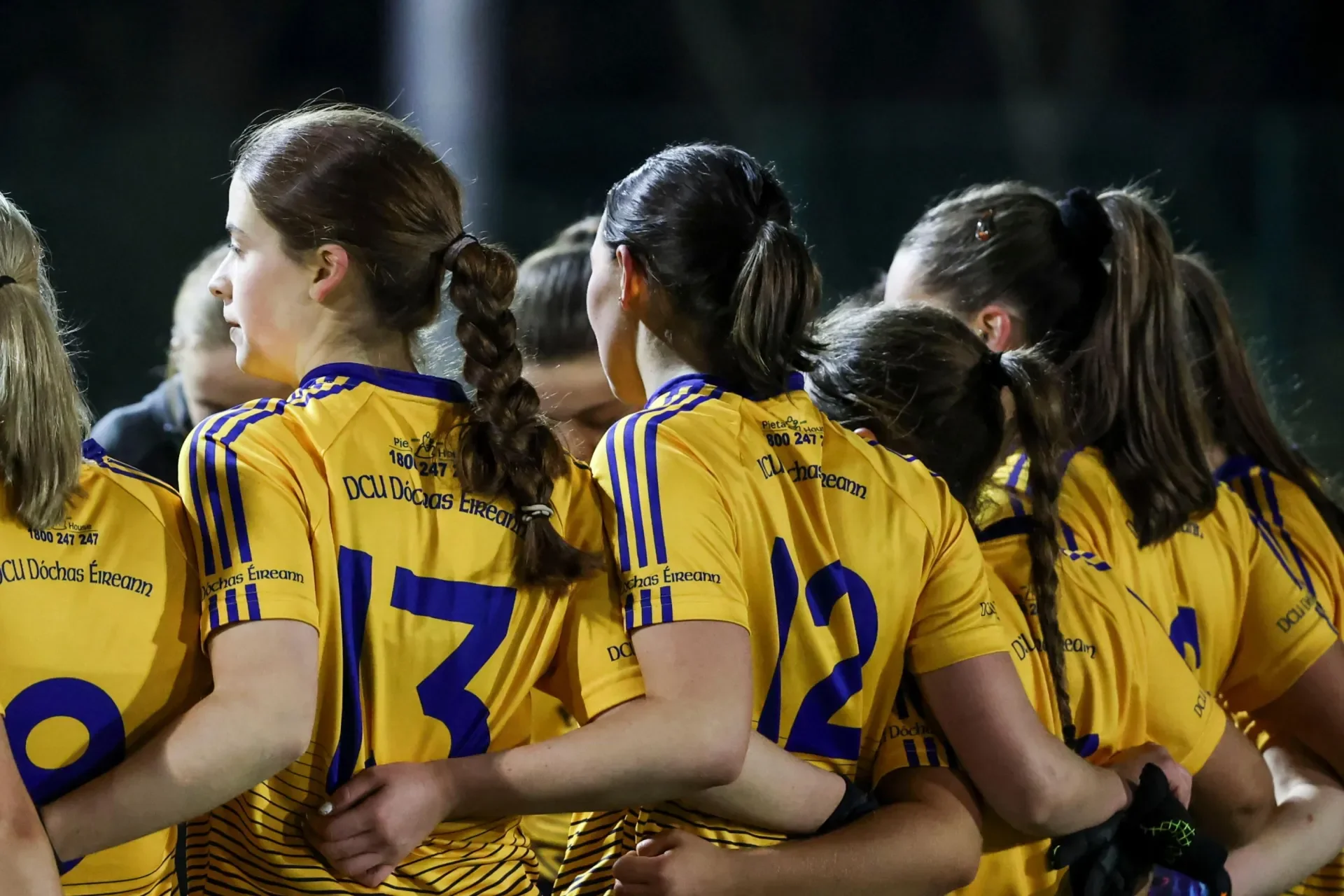 Shows DCU GAA players from behind, waist up with numbers visible on their jerseys