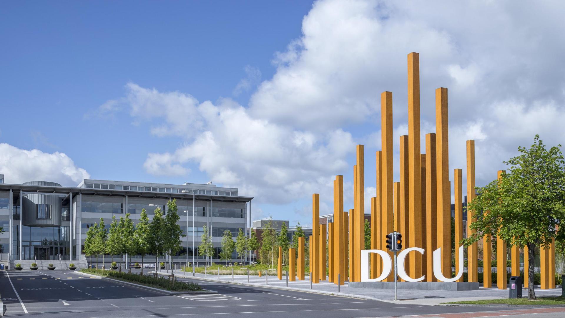 A view of DCU and it's surrounding modern buildings on a sunny day, featuring the DCU sign and the wooden sculpture next to it.