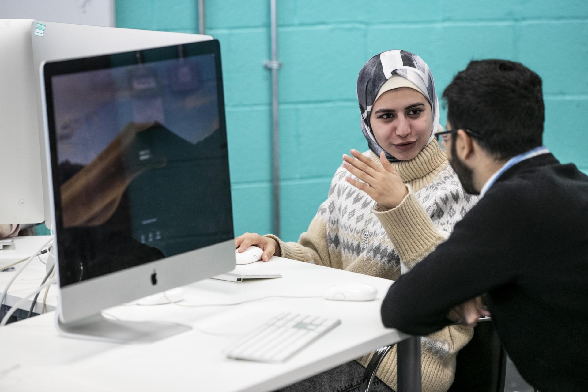 A female student with a head covering explaining to a male student in front of a computer