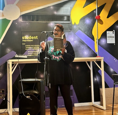 Poet reading their poetry at an open mic in front of a colorful wall