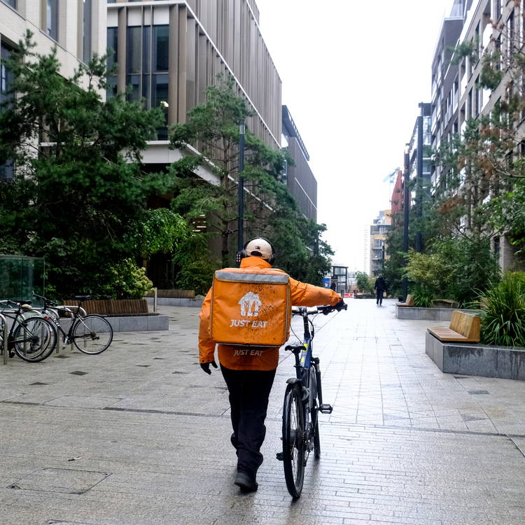 Delivery driver dressed in Orange walks with his bike through tall buildings lined with green trees