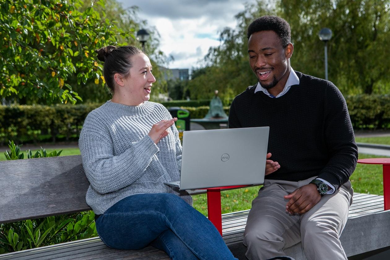 A pair of students sitting on a bench with a laptop on DCU's Glasnevin campus. 