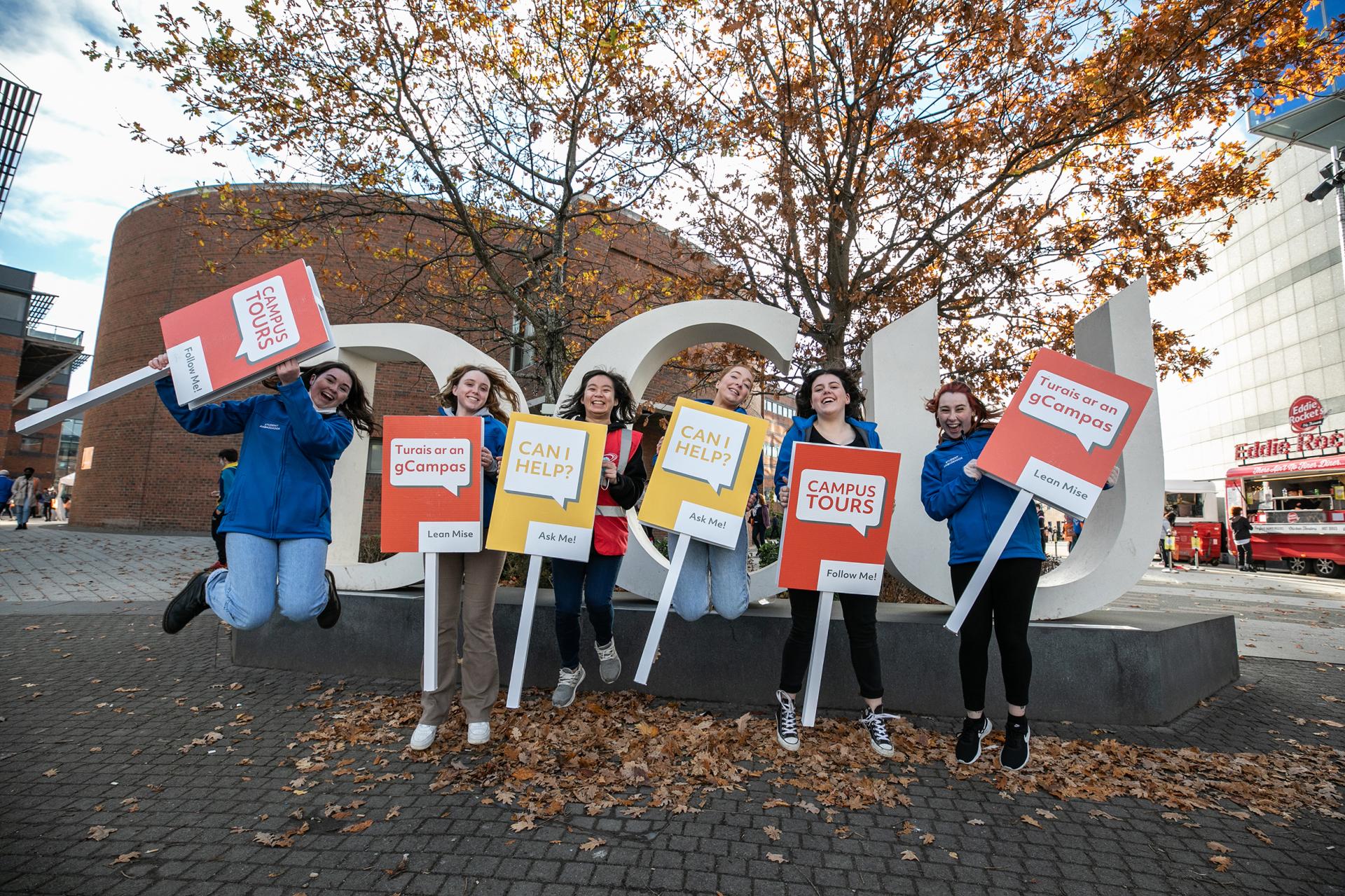 A group of Student Ambassadors are all jumping in front of the DCU Letters, holding signs encouraging people to ask questions and go on tours