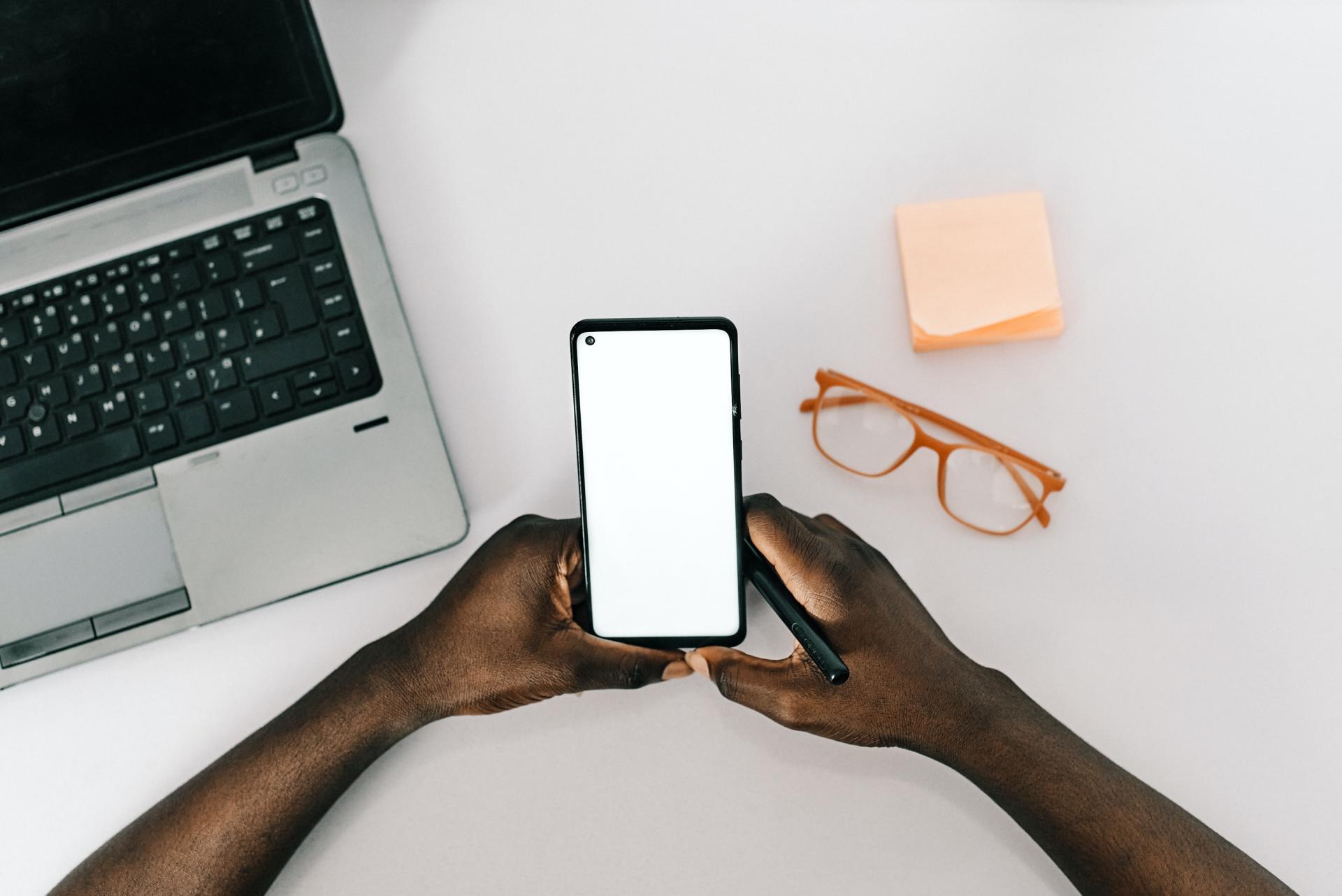 A smartphone in the hands of a person sitting in front of a laptop 