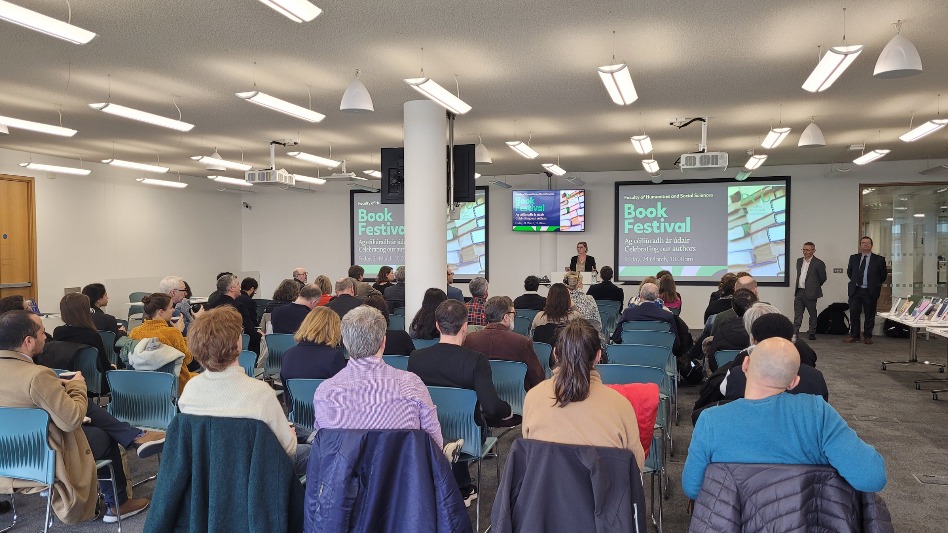 Shows a group of people seated in front of a presenter and a presentation screen that reads 'Book Festival.'