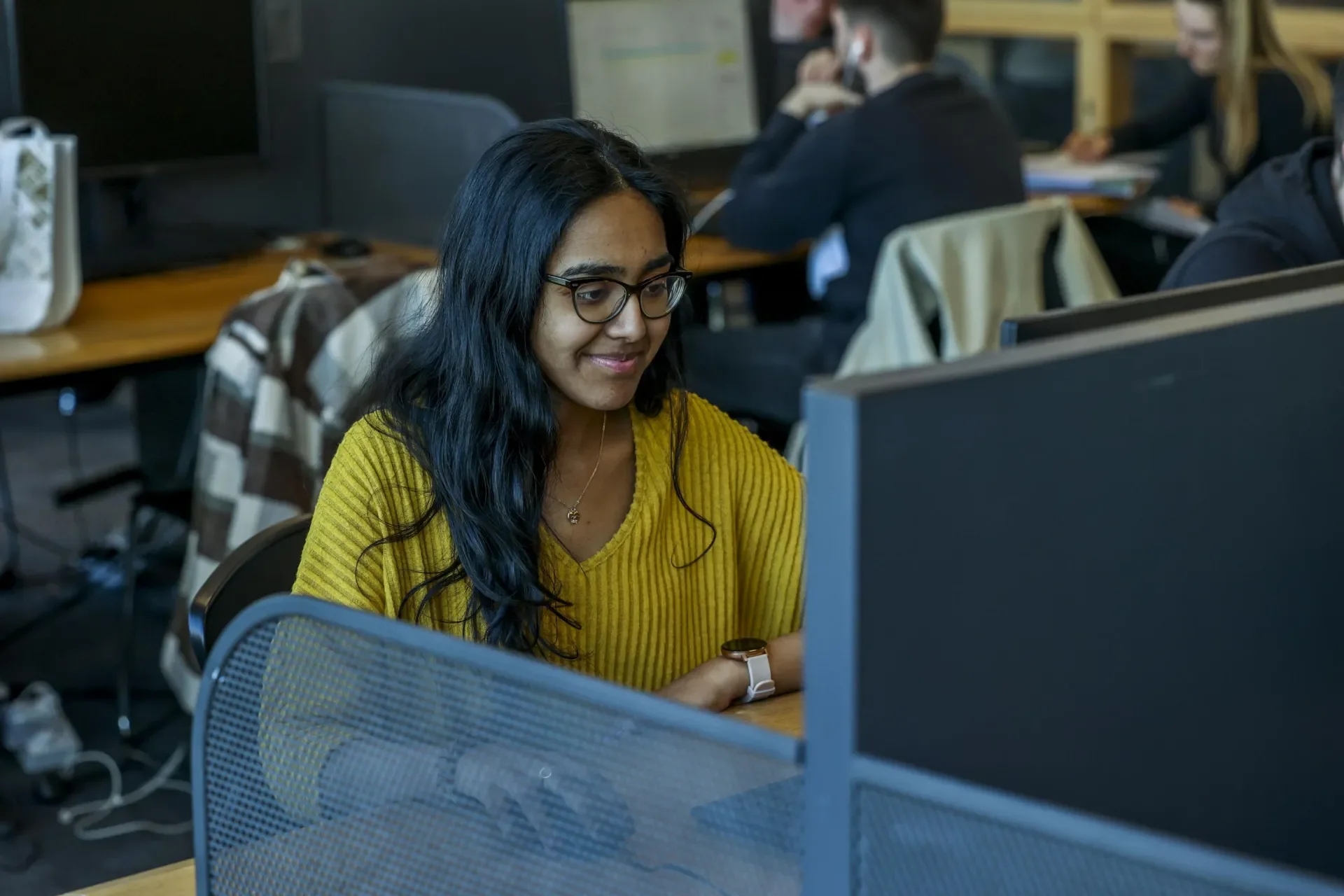 A woman sits in front of a computer. People are sitting in chairs at tables behind her.
