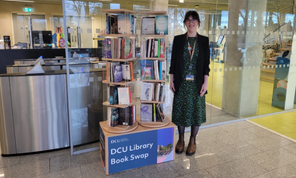 Shows staff member standing beside a carousel filled with books with DCU Library Book Swap written on the stand