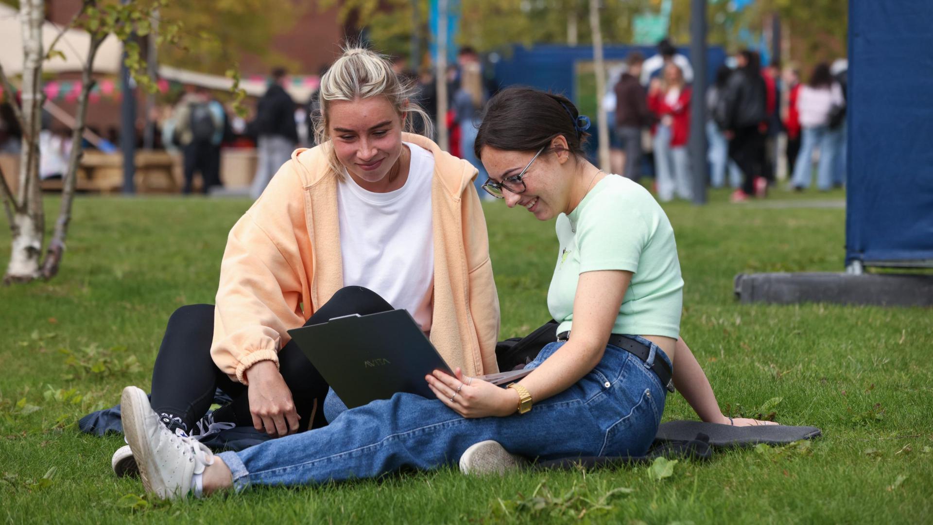 A photo of two young students sitting on some grass, reading from a laptop.