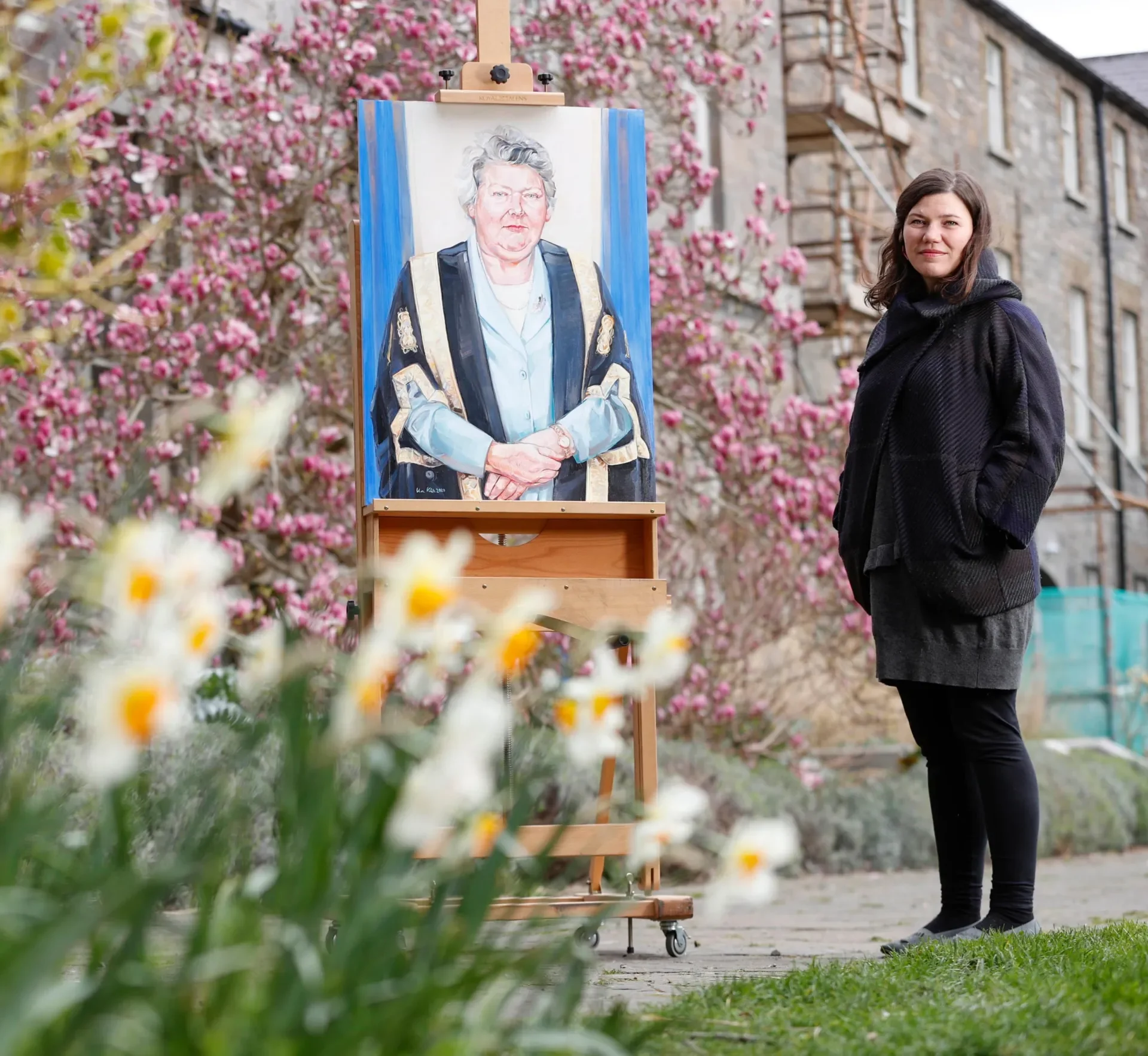 Artist Vera Klute, a woman with brown hair and black clothes stands beside an easel displaying her portrait of Ms Justice Mella Carroll