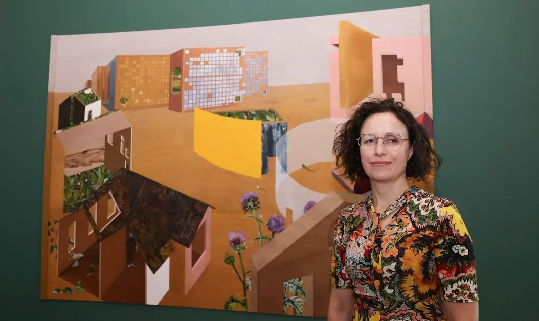 Artist Deirdre Frost, a women with brown curly hair and glasses stands in front of her artwork - Spreag. A painting which includes buildings from across the three campuses of DCU
