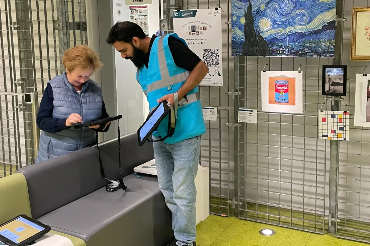 Person volunteering at Culture Night in high vis blue vest shows an older person an electronic tablet which explains the Art or AI exhibition